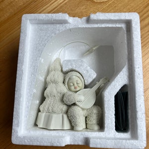 May include: A white porcelain figurine of a snow person playing a banjo, sitting on a snow-covered stump under a white tree with a star on top. The figurine is in a white styrofoam box.