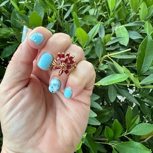 May include: A gold ring with a cluster of red gemstones in the centre. The ring is on a hand with light blue nail polish.