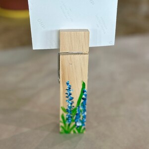 May include: A wooden clothespin with a painted design of blue flowers and green leaves. The clothespin is holding a white photo paper with the text "DNP" printed on it.