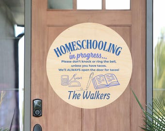 May include: A round wooden sign with the text "HOMESCHOOLING in progress..." in blue. The sign also includes the text "Please don't knock or ring the bell, unless you have tacos. We'll ALWAYS open the door for tacos!" and "The Walkers".