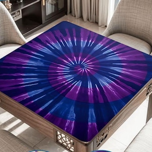 May include: A square table cover featuring a vibrant tie-dye pattern in shades of deep purple and royal blue. The design radiates from the center in a spiral motion, creating a bold, colorful display that covers the entire surface of the table.