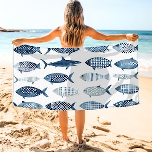 May include: A white beach towel featuring a pattern of various blue fish designs. The fish vary in shape and pattern, including polka dots, stripes, and geometric shapes. The towel is held up by a person on a sandy beach with the ocean in the background.