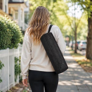 May include: A black cylindrical yoga mat bag with a long shoulder strap and a vertical zipper closure. The bag is worn over the shoulder, featuring a structured, elongated shape designed to hold a rolled mat securely for transport and storage.