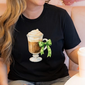 Może przedstawiać: Czarny t-shirt z akwarelową ilustracją napoju kawowego z bitą śmietaną i zieloną kokardą z koniczyną. Koszulka ma okrągły dekolt i krótkie rękawy.