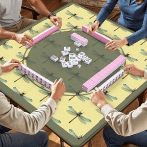 May include: A group of people playing Mahjong on a square table. The table has a green center and a yellow border with dragonfly patterns. Pink tile racks and white tiles are visible.