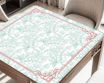 May include: A square wooden table with a white tablecloth featuring a delicate green floral pattern. The tablecloth has a thin, coral-colored border with decorative corner accents. The table is surrounded by chairs.