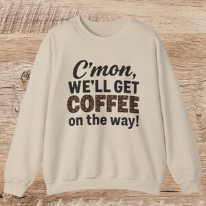 Puede incluir: Sudadera color crema con la frase "C'mon, WE'LL GET COFFEE on the way!" en letras negras y marrones. La palabra "COFFEE" está rellena con gráficos de granos de café. La sudadera tiene cuello redondo y mangas largas.