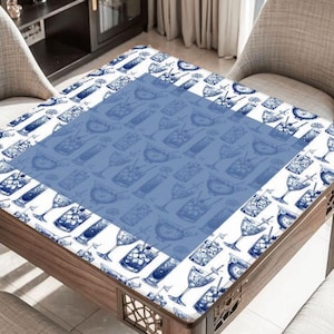 May include: A square wooden table with a white and blue cocktail-themed tablecloth. The tablecloth features a repeating pattern of cocktail glasses and bottles. The table is surrounded by two chairs.