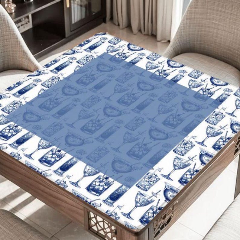 May include: A square wooden table with a white and blue cocktail-themed tablecloth. The tablecloth features a repeating pattern of cocktail glasses and bottles. The table is surrounded by two chairs.