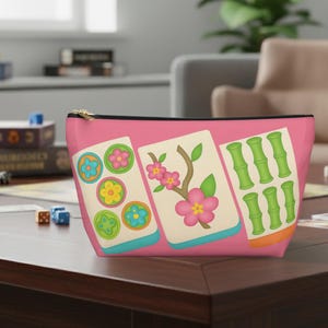 May include: A pink zippered cosmetic bag featuring colorful illustrations of Mahjong tiles. The bag displays floral and bamboo designs in green, pink, and yellow. The bag has a black zipper and a gold-colored zipper pull.