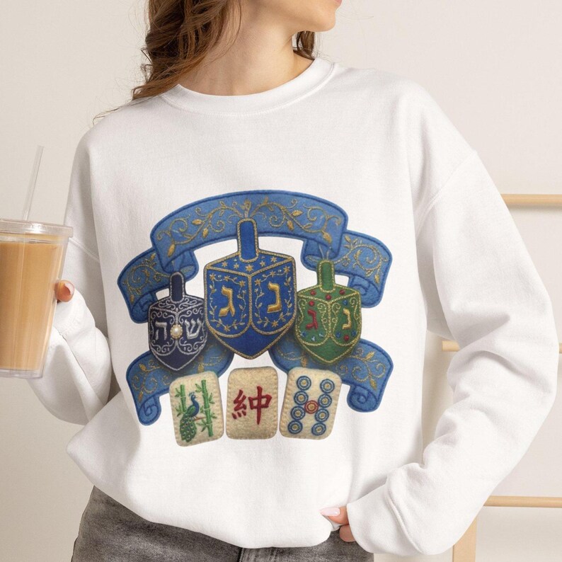 May include: A white crewneck sweatshirt featuring a graphic print of three dreidels in blue and green, set against a blue decorative ribbon banner with gold filigree. Below the dreidels are three mahjong tiles with a peacock, red text, and circles.