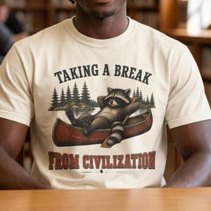 以下が含まれることがあります： クリーム色の半袖Tシャツ。湖に浮かぶ赤いカヌーの上でリラックスするアライグマと、背景に松の木が描かれたグラフィックが特徴です。上部にはTAKING A BREAK、下部にはFROM CIVILIZATIONという文字が暗い色でプリントされています。