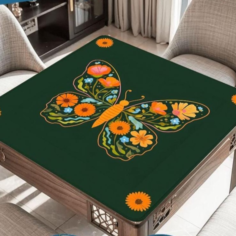 Puede incluir: Una cubierta de mesa cuadrada de color verde oscuro con un gran dise&ntilde;o de mariposa. La mariposa tiene patrones florales naranjas y amarillos en sus alas, con acentos azules. La mesa tiene un marco de madera y est&aacute; rodeada de sillas.