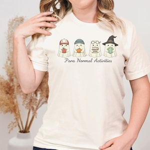 May include: Cream-colored t-shirt featuring a graphic of four cartoon ghosts, each wearing a different hat and holding an item. The text "Para Normal Activities" is printed below the ghosts. The ghosts are holding an apple, a pumpkin, books, and a globe.