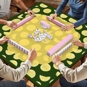 Pode incluir: Um jogo de Mahjong com jogadores em volta de uma mesa. A mesa tem uma superf&iacute;cie amarela com grandes c&iacute;rculos amarelos e uma borda verde com fatias de lim&atilde;o. Pe&ccedil;as e dados rosa s&atilde;o vis&iacute;veis.