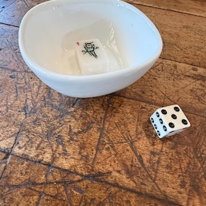 May include: A white ceramic bowl with a single Mahjong tile featuring a floral design and the number 1. A white and black dice with black dots is next to the bowl. The objects are on a wooden surface.