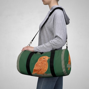 Puede incluir: Bolsa de lona verde con una ilustración detallada de un pájaro naranja. La bolsa tiene correas negras y una correa para el hombro negra. El diseño del pájaro está en la parte delantera y trasera de la bolsa. La bolsa es de forma cilíndrica.