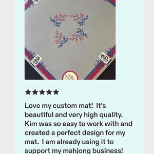 May include: A square light grey mahjong mat featuring a central pattern of four blue and red floral sprigs. The border displays a red and blue stripe design with a mahjong tile graphic in each corner. A circular sticker with the text Mahjong is visible.