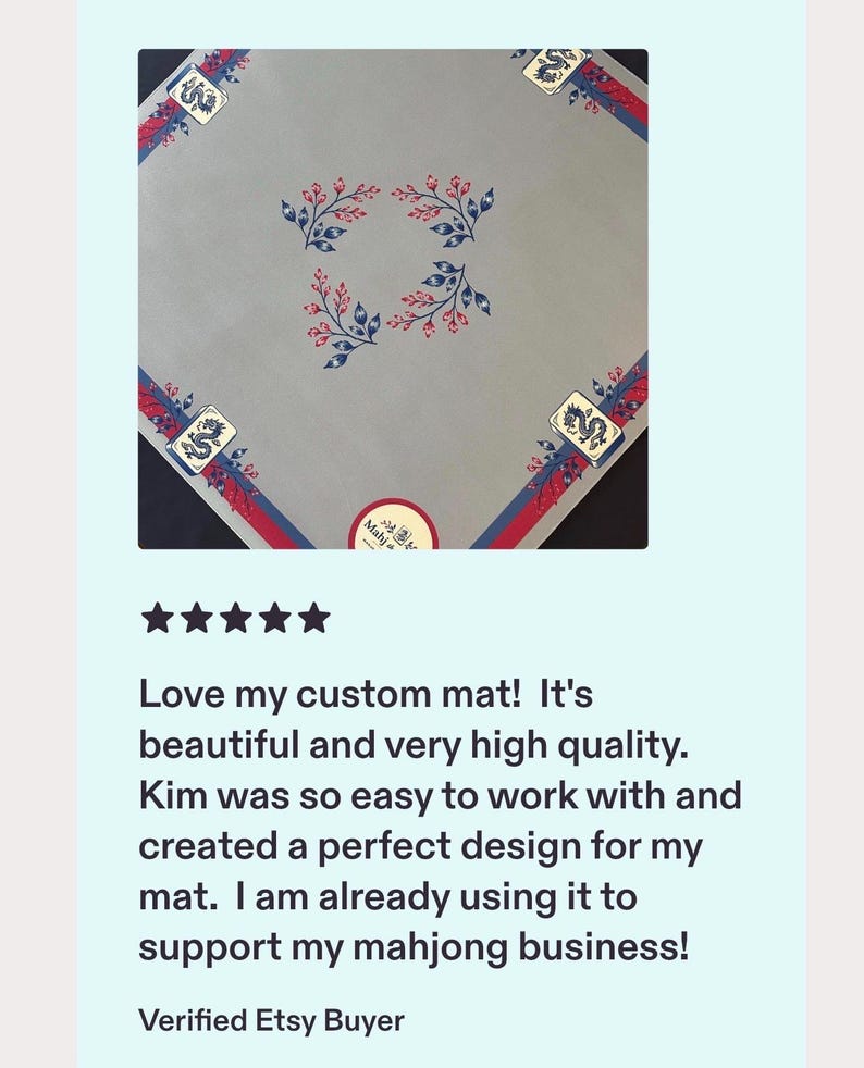 May include: A square light grey mahjong mat featuring a central pattern of four blue and red floral sprigs. The border displays a red and blue stripe design with a mahjong tile graphic in each corner. A circular sticker with the text Mahjong is visible.
