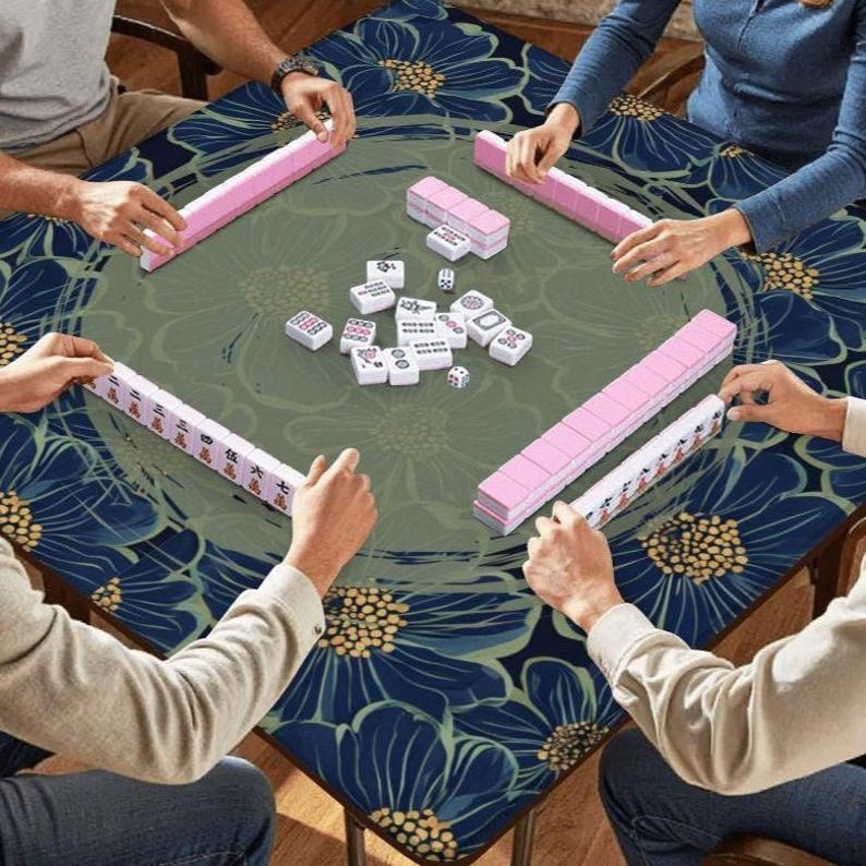 Op de afbeelding: Een vierkante tafel met een bloemmotief is klaar voor een spel Mahjong. Roze en witte tegels zijn gerangschikt, met spelers die tegelrekken vasthouden. De tafel heeft een groen midden en een blauwe bloemenrand.