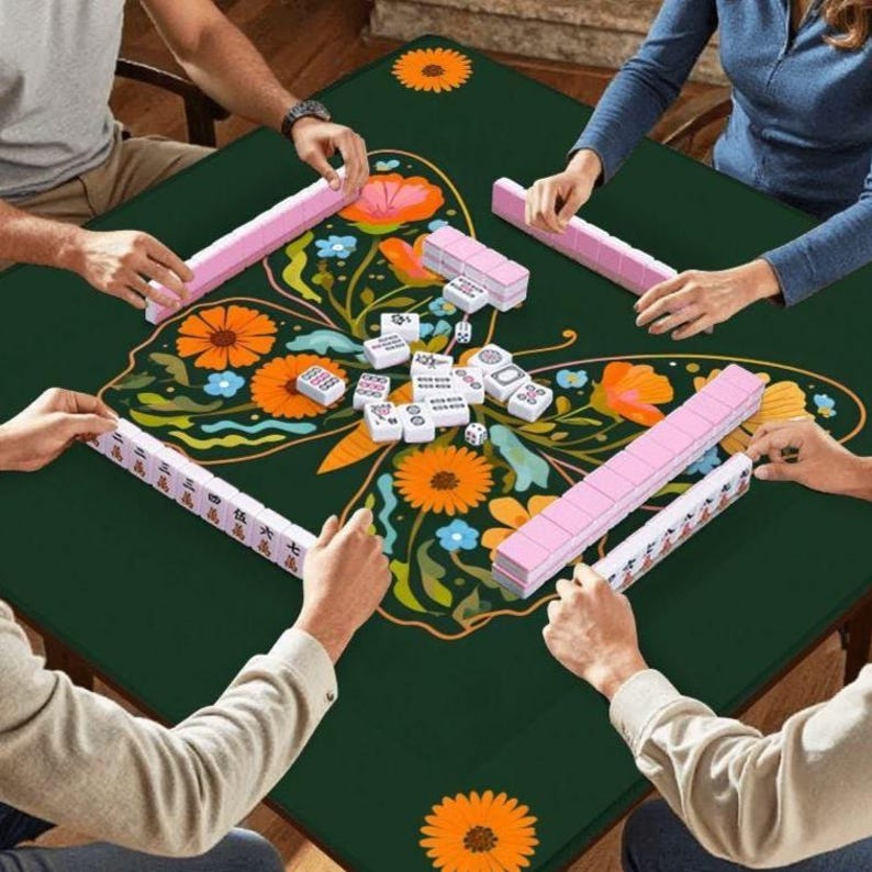 Puede incluir: Una partida de mahjong en curso sobre una mesa verde oscuro. La mesa presenta un dise&ntilde;o de mariposa colorido con flores naranjas y rosas. Las fichas de mahjong rosas est&aacute;n dispuestas alrededor de la mesa, y los jugadores sostienen sus fichas.