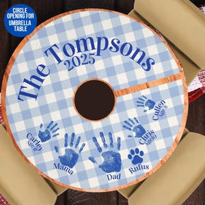 May include: Blue and white checkered table cover with the text "The Tompsons 2025" in blue. The cover has handprints of family members and a paw print of a dog. The names and ages of the family members are written below their handprints.