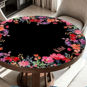 May include: A round table cover featuring a black background with a vibrant floral border of red, pink, orange, and purple flowers and green foliage. The name KIM is printed in a light orange font on the right side of the floral pattern.