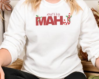 May include: White sweatshirt with the text "merry MAH" in red letters. The "A" is decorated with holly leaves, and the "J" is a candy cane. The sweatshirt is long-sleeved and has a crew neck.