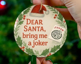 May include: A round ceramic Christmas ornament featuring a red Santa hat at the top and a green holly leaf border. The text reads Dear SANTA, bring me a joker, accompanied by a small illustration of a playing card labeled JOKER.