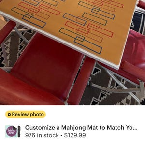 May include: A custom mahjong mat with a tan surface and a white border. The mat features a geometric design of red and blue rectangles. Red chairs surround the table.