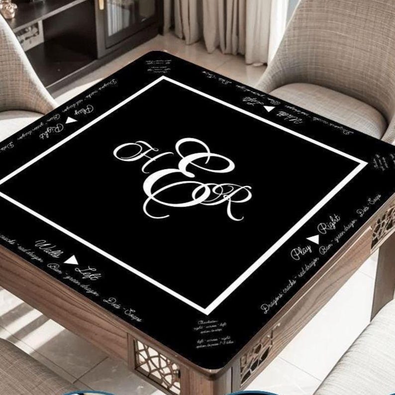 May include: A square black mahjong table mat featuring a white border and a central white monogram of the letters H and E. The mat includes white text printed along the edges with directional instructions and game terminology in English.