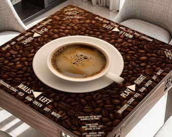 May include: A square table covered in a coffee bean pattern featuring Mahjong game instructions printed in white text. A white ceramic cup filled with dark coffee sits on a matching saucer in the center of the table surface.