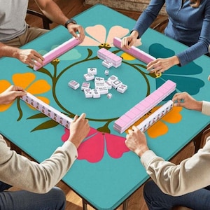 May include: A square teal game table with a floral design is set for a game of Mahjong. Pink tiles and dice are scattered on the table. Players are holding pink tile racks and game tiles.