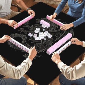 May include: A black Mahjong table with a white dragon design is surrounded by players. Pink tile racks and white tiles with symbols are visible. The game is in progress, with players holding tiles.