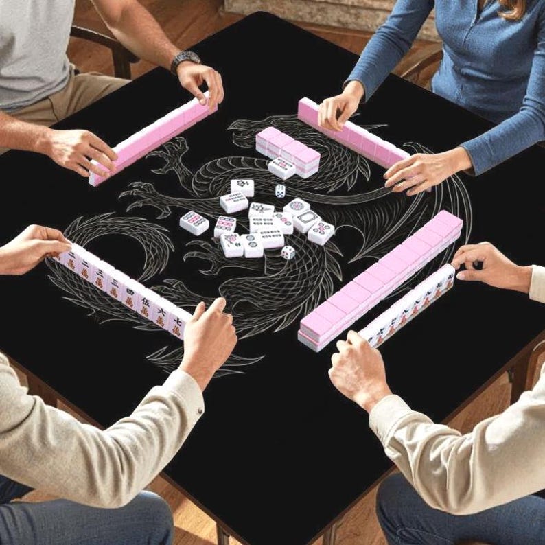 May include: A black Mahjong table with a white dragon design is surrounded by players. Pink tile racks and white tiles with symbols are visible. The game is in progress, with players holding tiles.