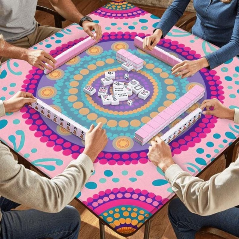 Puede incluir: Una mesa de Mahjong cuadrada con un colorido dise&ntilde;o de mandala en rosa, morado y turquesa. El juego est&aacute; en curso, con jugadores sosteniendo estantes de fichas rosas y fichas blancas. La mesa est&aacute; rodeada de personas.