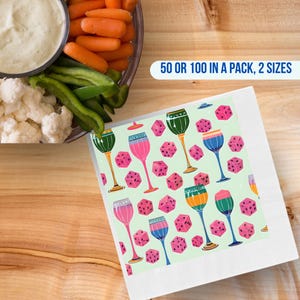 Könnte beinhalten: Weiße Papierservietten mit einem bunten Muster aus Weingläsern und rosa Würfeln auf hellgrünem Hintergrund. Die Servietten liegen auf einer Holzoberfläche, mit einer Schüssel mit Dip und Gemüse im Hintergrund. Text auf dem Bild: 50 ODER 100 IN EINER PACKUNG, 2 GRÖSSEN.