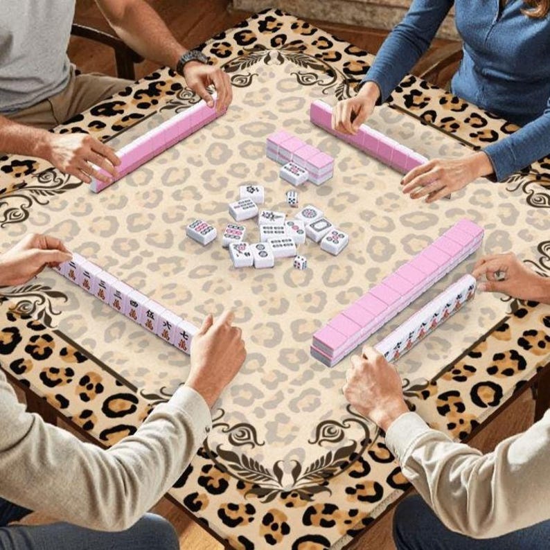 Pode incluir: Um grupo de pessoas jogando Mahjong em uma mesa com um design de estampa de leopardo. As pe&ccedil;as do jogo s&atilde;o rosa e brancas. A mesa tem um fundo bege com um padr&atilde;o de estampa de leopardo e uma borda decorativa.