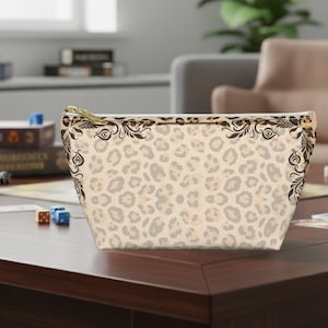 May include: A beige cosmetic bag with a leopard print pattern. The bag features a gold zipper and decorative black floral accents along the top edges. The bag is sitting on a dark brown wooden surface.