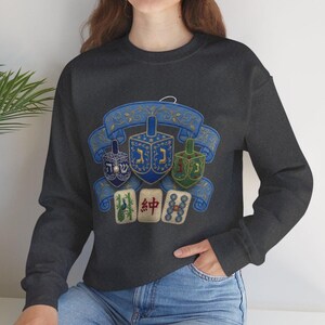 May include: A dark gray long-sleeve sweatshirt featuring a graphic print of three dreidels in blue, dark blue, and green, positioned in front of a blue decorative ribbon banner, with three mahjong tiles displayed below the dreidels.