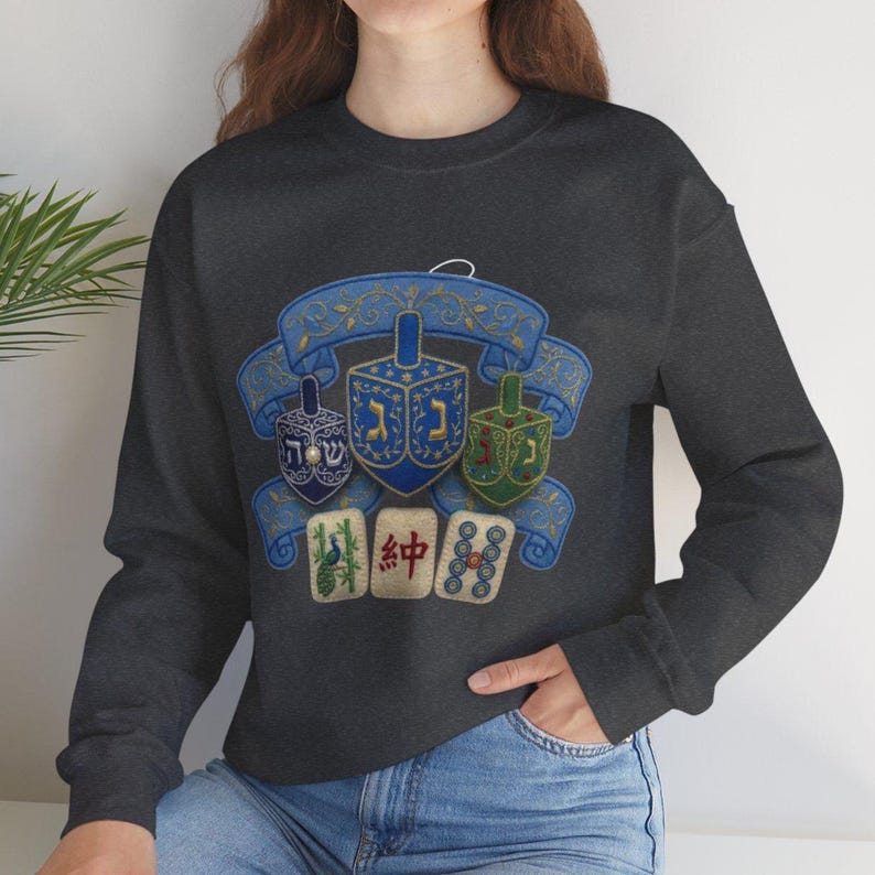 May include: A dark gray long-sleeve sweatshirt featuring a graphic print of three dreidels in blue, dark blue, and green, positioned in front of a blue decorative ribbon banner, with three mahjong tiles displayed below the dreidels.