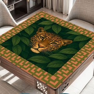 May include: Square wooden table with a decorative top featuring a leopard's face surrounded by green leaves. The table's border has an orange and green geometric pattern. The table is set in a room with two gray chairs.
