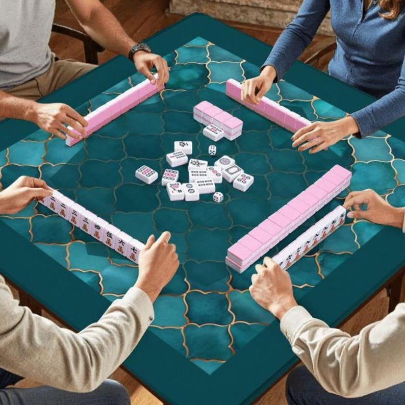 May include: A square mahjong table featuring a teal surface with a gold quatrefoil pattern. Four players are seated around the table, each with a rack of pink-backed tiles. White mahjong tiles with black and red characters are arranged in the center.