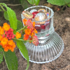 May include: A clear glass candle holder with a fluted design. The holder is filled with iridescent glass beads in shades of red, gold, and silver.