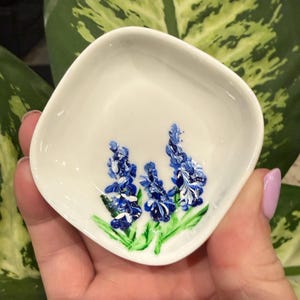 May include: A small, white, square-shaped ceramic dish with rounded corners. The center of the dish features a hand-painted design of three blue flower stalks with green leaves, rendered in a textured, raised paint style against the smooth surface.