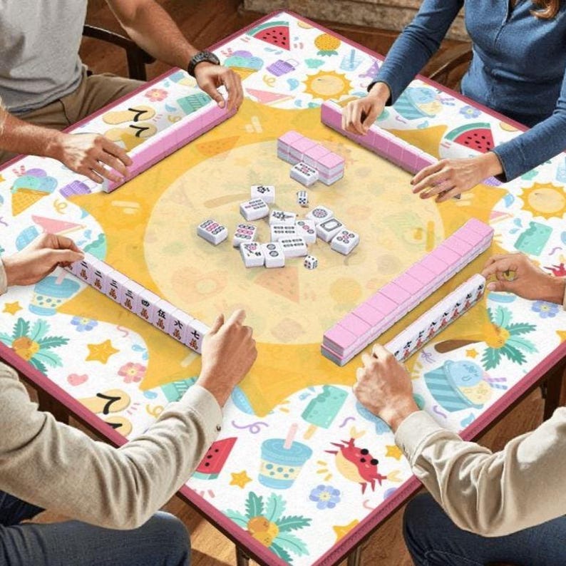 May include: A square mahjong table featuring a white surface with a large yellow sun graphic and colorful summer-themed icons including watermelons, ice cream, and crabs. Four players are seated around the table with rows of white mahjong tiles.