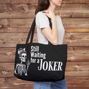 May include: Black tote bag with the text "Still Waiting for a JOKER" in white. The bag features a white illustration of a skeleton with a bow in its hair. The bag has black straps.