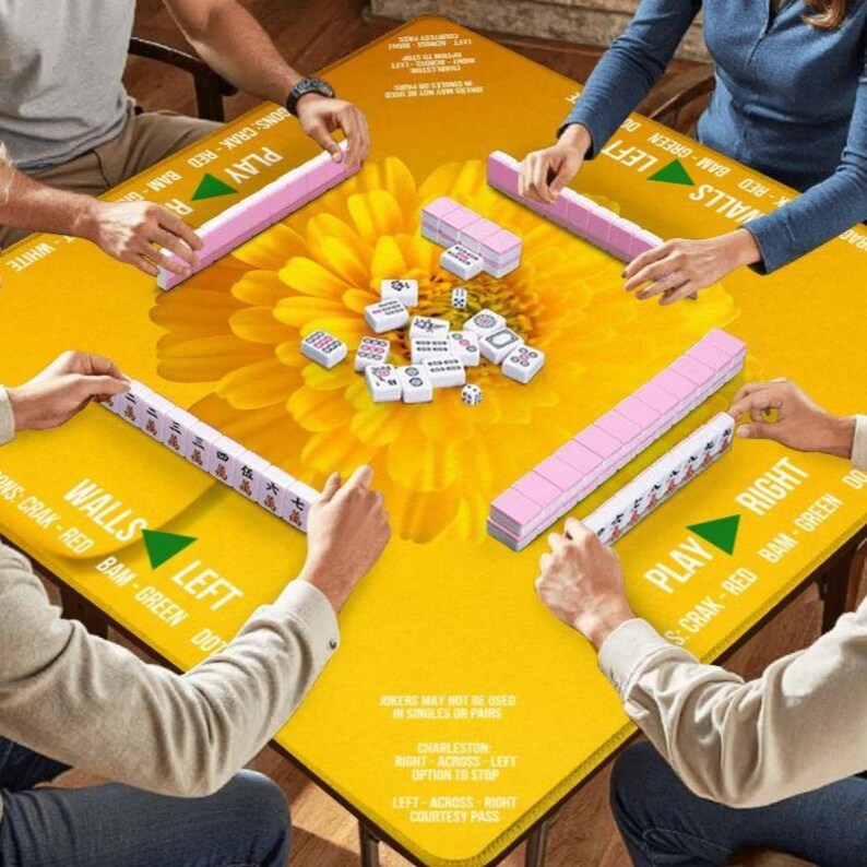 Puede incluir: Una mesa de Mahjong amarilla con un gran dise&ntilde;o floral est&aacute; rodeada de jugadores. Las fichas rosas y blancas est&aacute;n dispuestas sobre la mesa. La mesa tiene el texto "Walls Left" y "Play Right".