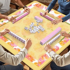 May include: A square yellow Mahjong table with a floral design and pink accents. The table is surrounded by players holding pink tile racks and playing tiles. Dice and additional tiles are in the center.