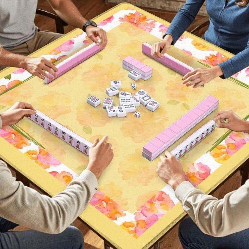 May include: A square yellow Mahjong table with a floral design and pink accents. The table is surrounded by players holding pink tile racks and playing tiles. Dice and additional tiles are in the center.